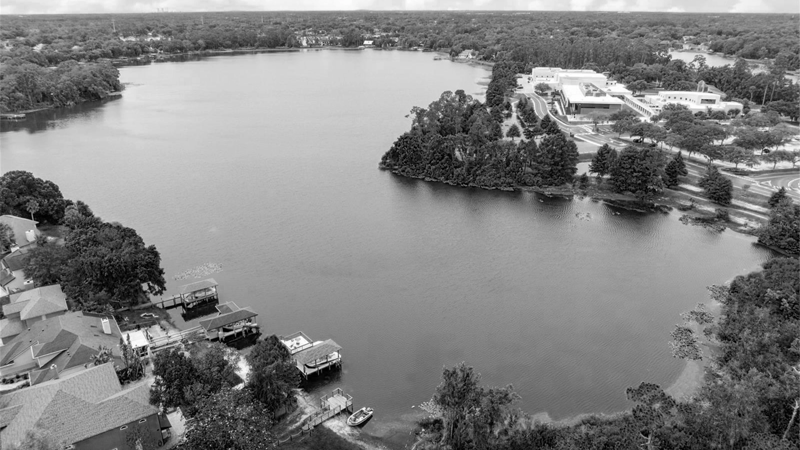 Black and white aerial drone photograph of Lake Burkett in Central Florida, showing a wide lake surrounded by waterfront homes with private boat docks, lush green trees, and large commercial or institutional buildings on the opposite shore.