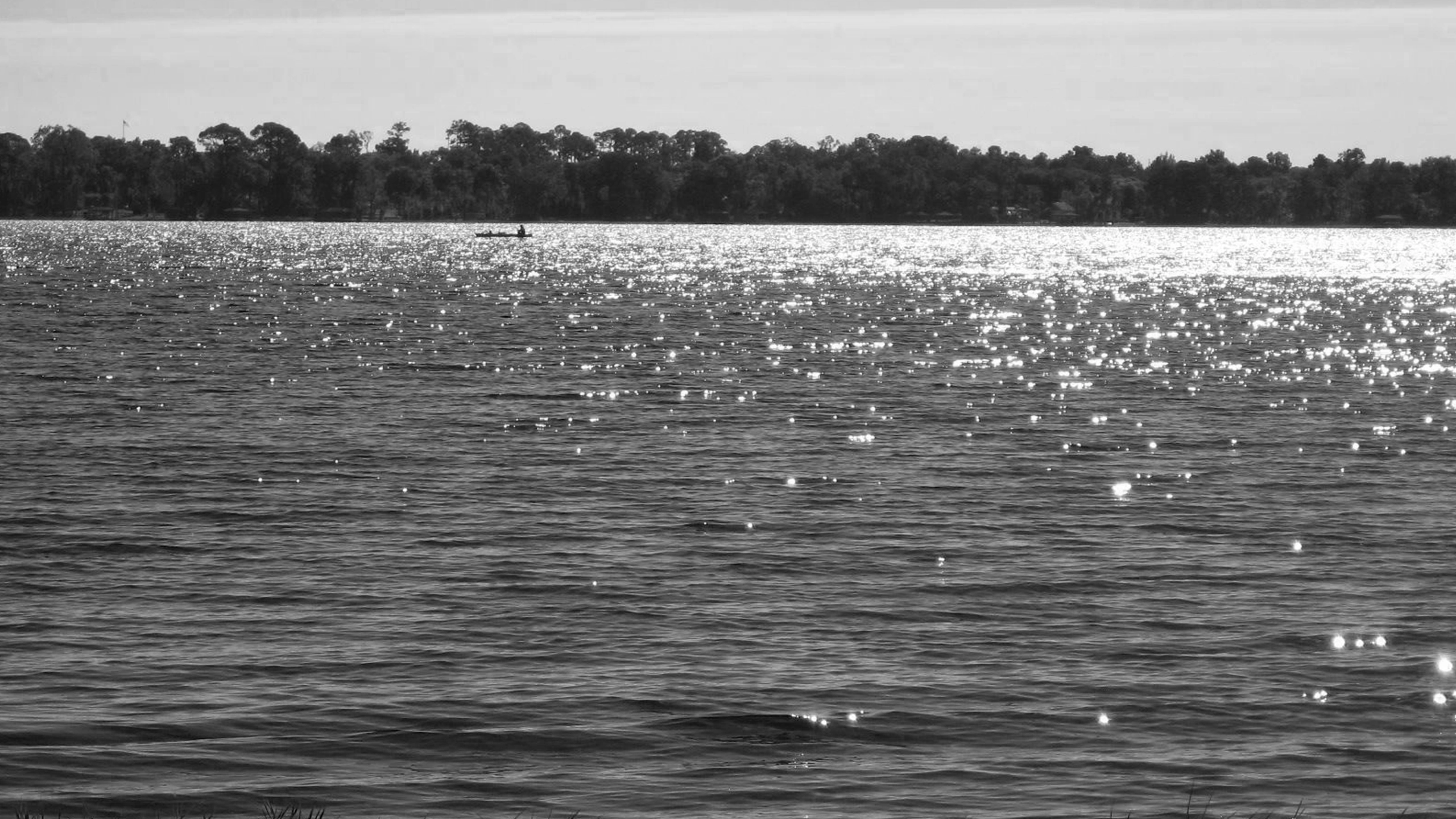 A black and white landscape photograph of Lake Sheen, featuring bright sunlight creating a brilliant shimmering glare across the rippling surface of the water, with a small boat silhouetted in the distance against a heavily wooded far shoreline.